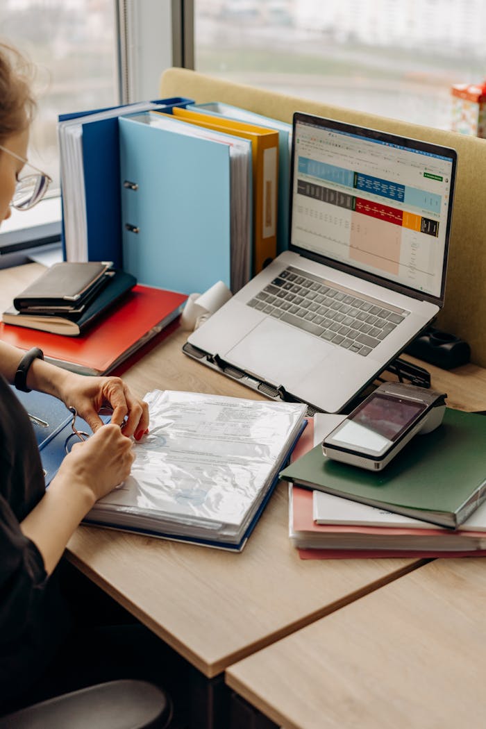 A woman organizing documents at a desk with binders and a laptop in an office setting.